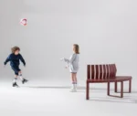 Urbantime Liberty.080 curved outdoor bench shown in a playful lifestyle setting with children playing football nearby.