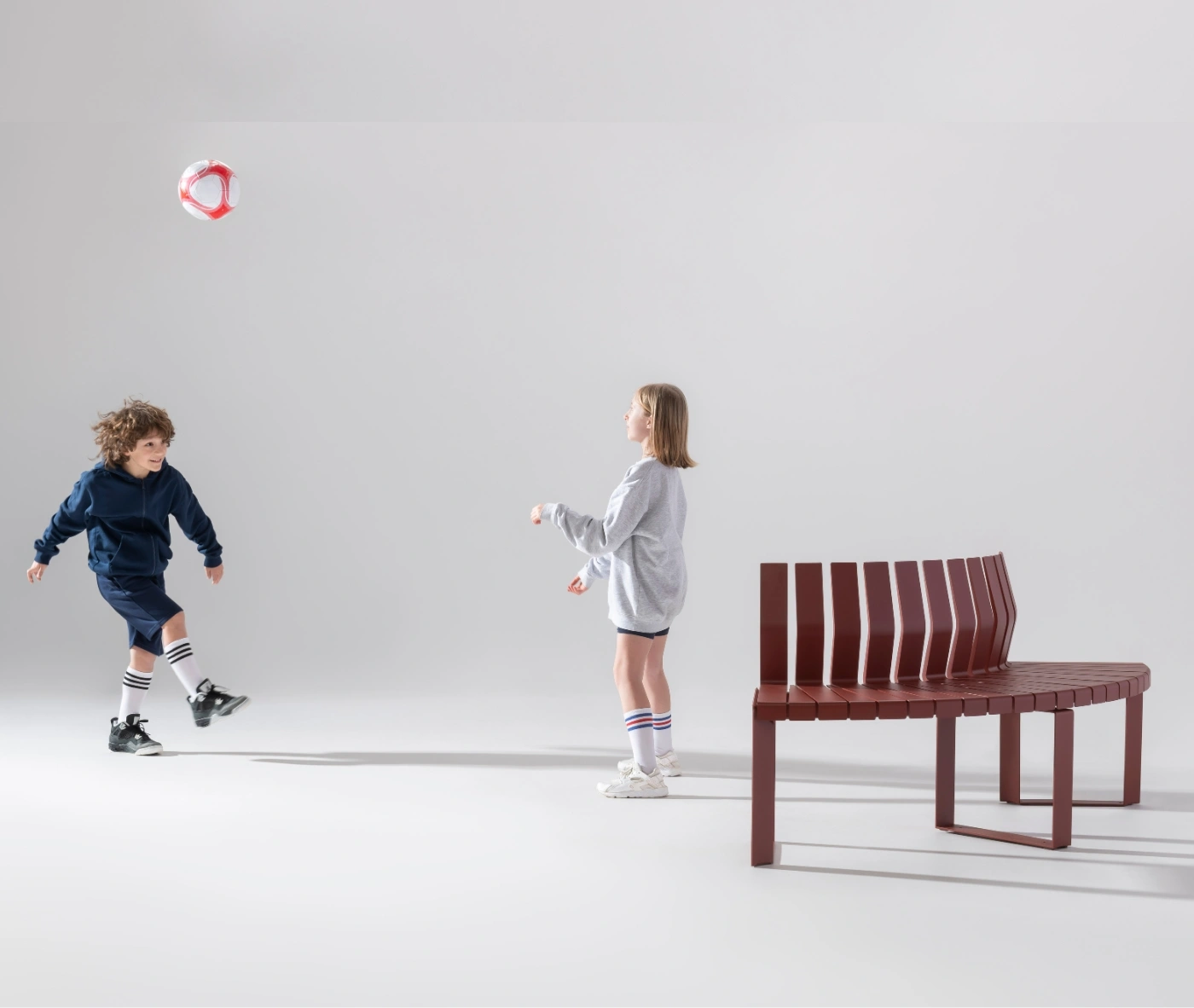Urbantime Liberty.080 curved outdoor bench shown in a playful lifestyle setting with children playing football nearby.