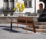 Urbantime Liberty.080 outdoor slat bench in front-left view set in an urban plaza.